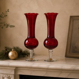 May include: A pair of tall, deep red glass vases with flared, scalloped rims and bulbous bodies. Each vase is supported by a clear, textured glass stem and a circular base, creating a distinct contrast between the vibrant red and clear glass.
