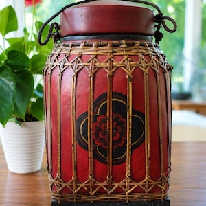 May include: A cylindrical red container with a lid, featuring a black floral motif and a woven bamboo lattice overlay. The base is a solid black square block with carved flower details, and a dark cord handle is attached to the top lid.