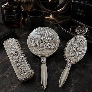 May include: A silver vanity set consisting of a rectangular clothes brush, a round hand mirror, and an oval hairbrush. Each piece features an intricate embossed scene of figures in a landscape. The handles are smooth, polished silver metal.