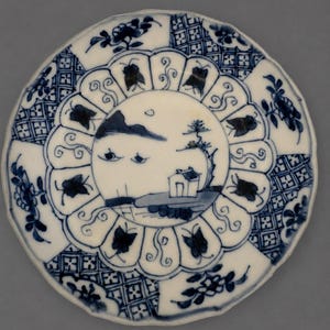 May include: A round ceramic plate featuring a blue and white hand-painted design. The center depicts a landscape with a small building, a tree, and mountains. A scalloped border surrounds the center, decorated with butterfly motifs and floral patterns.