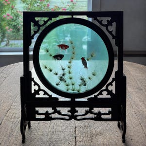 May include: A square dark wood frame with a circular center containing a translucent light blue panel. The panel features an embroidered or painted scene of three small fish in red and black, surrounded by thin, spiky green aquatic plants.