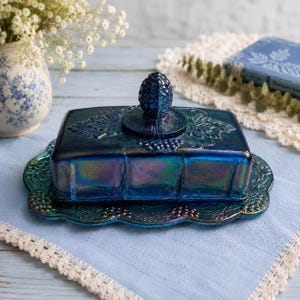 May include: A rectangular iridescent blue glass butter dish with a matching lid. The lid features a central pineapple-shaped finial and embossed grape leaf patterns. The base has a scalloped edge with matching embossed grape and leaf details.