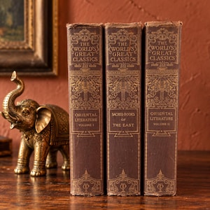 May include: Three brown hardcover books titled The World's Great Classics, including Oriental Literature Volume I, Sacred Books of the East, and Oriental Literature Volume II, standing beside a small metallic gold elephant figurine.