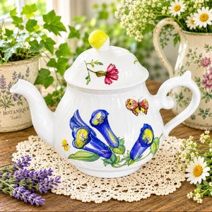 Op de afbeelding: Een witte keramische theepot met een handgeschilderd ontwerp van felblauwe klokvormige bloemen, een kleine vlinder en een gele bij. Het deksel is voorzien van een gele knop en de pot heeft een subtiele geribbelde textuur over het gehele oppervlak.
