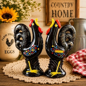 May include: Two black ceramic rooster figurines with red combs, yellow beaks, and yellow feet. The roosters have colorful accents and are displayed on a decorative doily. A jar with the words "Farm Fresh Eggs" is in the background.