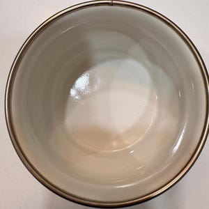 May include: A white bowl with a metallic rim. The bowl has a smooth, glossy interior and a slightly off-white colour. The rim is a thin band of metal, adding a touch of elegance to the simple design. The bowl is empty.