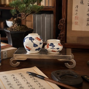 May include: Two white ceramic vessels with blue pine tree and orange bat motifs sit on a rectangular silver-toned metal stand. The vessels have rounded shapes and are decorated with traditional patterns, positioned centrally on the elevated display base.