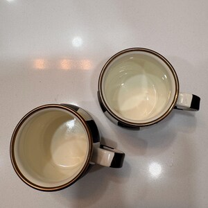 May include: Two ceramic mugs with a black and white checkered pattern. The mugs have a cream-colored interior and a gold-colored rim. The handles are black and white. The mugs are on a white surface.