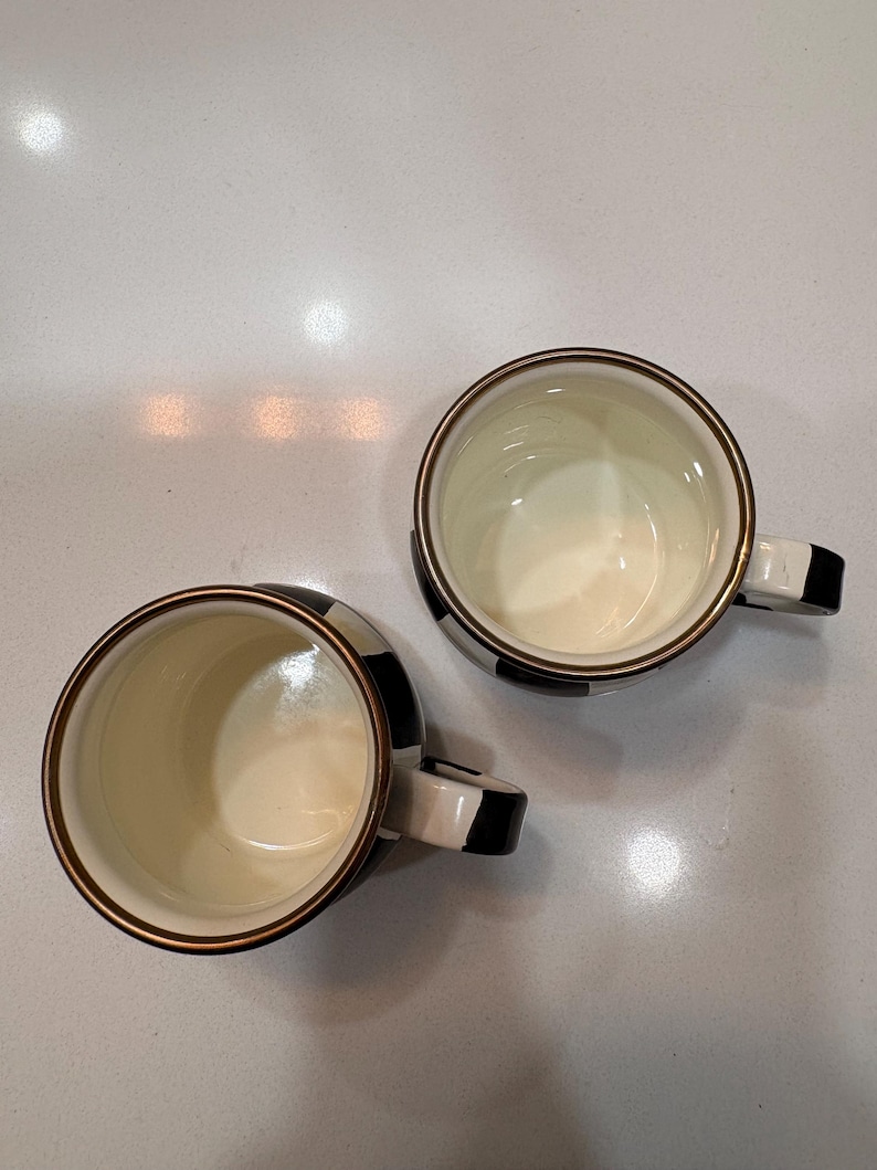 May include: Two ceramic mugs with a black and white checkered pattern. The mugs have a cream-colored interior and a gold-colored rim. The handles are black and white. The mugs are on a white surface.