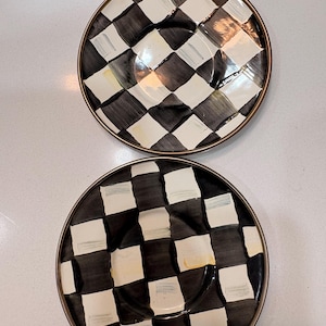 May include: Two decorative plates with a black and white checkered pattern. The plates have a gold-colored rim and a central indentation. The design features alternating black and white squares, creating a bold visual effect.