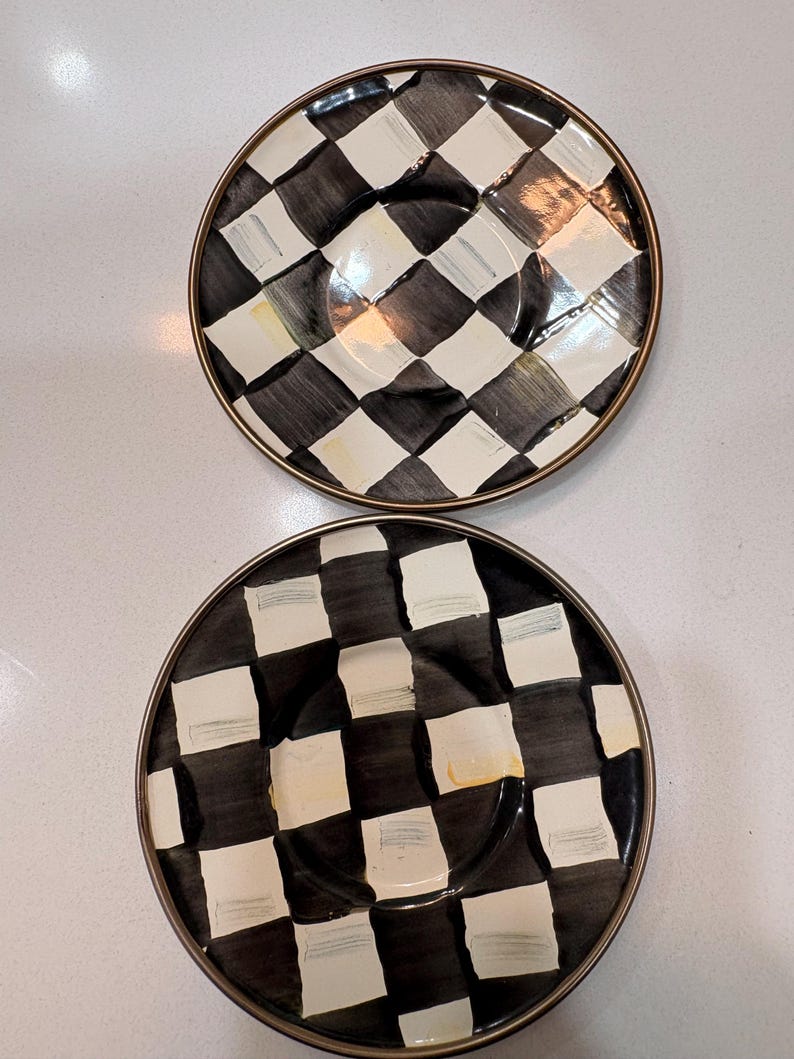 May include: Two decorative plates with a black and white checkered pattern. The plates have a gold-colored rim and a central indentation. The design features alternating black and white squares, creating a bold visual effect.