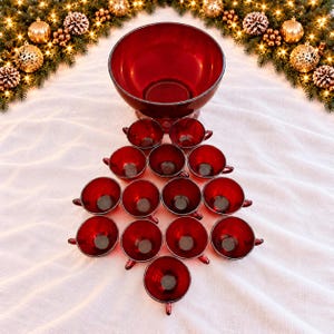 May include: A large, deep red glass punch bowl positioned above a set of twelve matching red glass punch cups arranged in a triangular, tree-like formation. The glassware features a smooth, translucent finish and a consistent deep red hue.