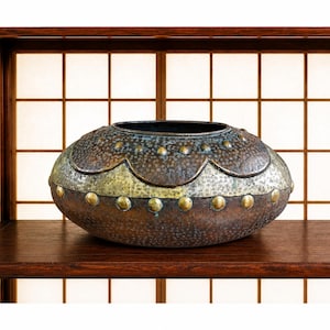 May include: A wide, rounded metal bowl with a dark, textured brown finish. The upper section features a scalloped border with small, raised brass-colored studs, while a central band displays a lighter, metallic, grid-like pattern.
