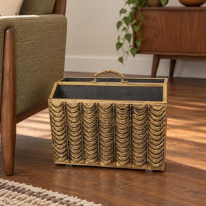 May include: A rectangular magazine rack with a metallic gold finish and a textured, repeating scalloped pattern on the exterior. The open-top container features a central metal handle on the top edge and a dark interior lining for storage.