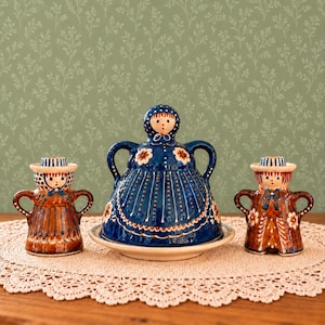 May include: A ceramic butter dish with a lid shaped like a person in a blue dress, flanked by two matching ceramic candle holders in brown, each featuring a person with a hat, painted floral details, and two side handles on each piece.