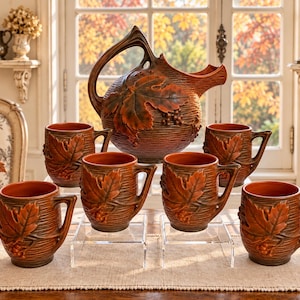 May include: A ceramic pitcher and six matching mugs featuring a raised relief design of maple leaves and grape clusters. The set has a deep brown and orange glaze with a textured, horizontal ribbed finish on the exterior and smooth interiors.