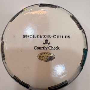 May include: A round decorative container with a white interior and a black and white checkered rim. The text "MACKENZIE-CHILDS" and "Courtly Check" are printed on the lid. A small oval sticker reads "MADE IN ROMANIA".