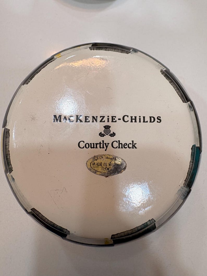 May include: A round decorative container with a white interior and a black and white checkered rim. The text "MACKENZIE-CHILDS" and "Courtly Check" are printed on the lid. A small oval sticker reads "MADE IN ROMANIA".