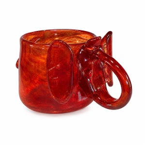 May include: A vibrant red glass mug with an elephant design. The mug features a large, curved handle and two large, flat ear-like elements on the sides. The glass has a marbled effect with shades of red and orange.