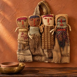 May include: A wall hanging featuring four stylized fabric dolls crafted from woven textiles in shades of brown, tan, and cream. Each doll has embroidered facial features in red and black thread, with the rightmost doll displaying a textured beard.