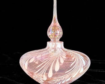 May include: A translucent pink glass perfume bottle with an iridescent finish and a wide, rounded base. The bottle features a delicate white feather-like pattern swirling across its surface and includes a tall, slender, matching glass stopper.