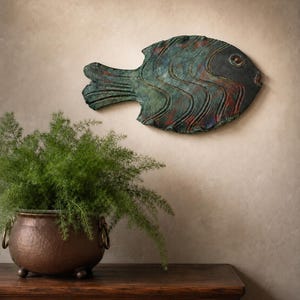 May include: A wall-mounted fish sculpture featuring a weathered patina finish in shades of teal, green, and copper. The fish has a dark, solid-colored head, a textured body with wavy vertical ridges, and a wide, flat tail fin design.