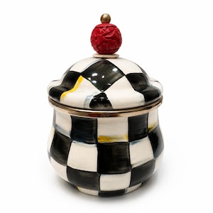 May include: A ceramic jar with a lid, decorated with a black and white chequered pattern. It has gold trim and a red, carved, spherical finial on the lid, with a small gold accent. The jar is set against a white background.