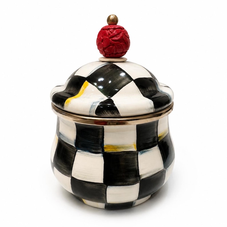 May include: A ceramic jar with a lid, decorated with a black and white chequered pattern. It has gold trim and a red, carved, spherical finial on the lid, with a small gold accent. The jar is set against a white background.