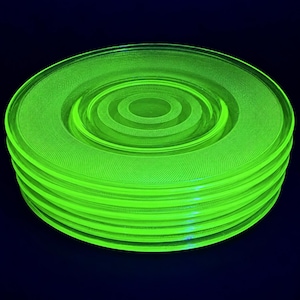 May include: A stack of five circular glass plates glowing with a vibrant neon green hue. Each plate features a concentric ring pattern with a textured, stippled surface detail on the rim and center, creating a uniform appearance across the set.