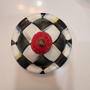 May include: A decorative ceramic lid with a red, textured knob. The lid features a black and white chequered pattern with accents of yellow and blue. The knob has a brass-coloured base.