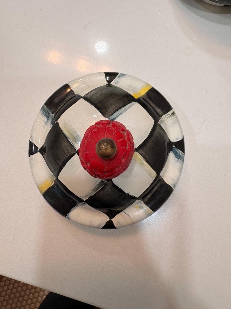 May include: A decorative ceramic lid with a red, textured knob. The lid features a black and white chequered pattern with accents of yellow and blue. The knob has a brass-coloured base.