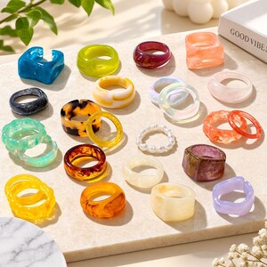 May include: A collection of assorted resin and plastic rings in various colors, shapes, and textures, including translucent, marbled, and solid finishes. The rings are arranged on a flat surface, featuring shades of blue, yellow, green, orange, purple, and white, with one ring made of small white beads.