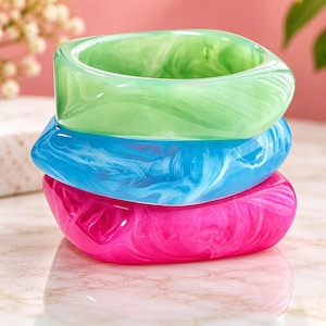 May include: A stack of three chunky bangle bracelets with a smooth, polished finish. The top bangle is light green, the middle bangle is bright blue, and the bottom bangle is vibrant pink. Each bracelet features a swirling, marbled pattern.