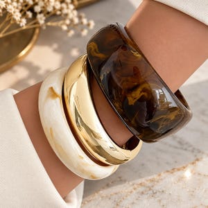 May include: A set of three bangles worn together. One bangle is a wide, dark brown resin with a swirling pattern. The second is a smooth, polished gold-tone metal. The third is a wide, off-white resin with subtle, light brown marbled veining.