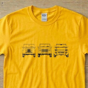 Peut inclure: Un t-shirt jaune vif à manches courtes orné d'un dessin au trait noir représentant trois générations différentes de véhicules Land Rover disposées horizontalement au centre de la poitrine. Le t-shirt possède un col rond et une coupe standard.