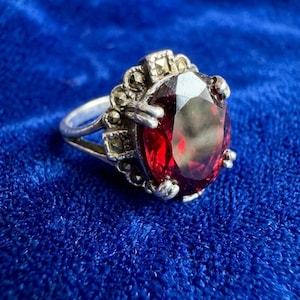 Puede incluir: Un anillo de plata con una gran piedra preciosa roja oscura de talla ovalada. El anillo presenta detalles intrincados alrededor de la piedra preciosa y en la banda. El engaste incluye pequeños acentos brillantes. El anillo se muestra sobre terciopelo azul.