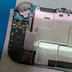 K&ouml;nnte beinhalten: Ein ge&ouml;ffnetes teal- und grau-farbenes Handheld-Spielger&auml;t, das interne Komponenten zeigt. Der obere linke Eckbereich des Ger&auml;ts zeigt einen abgenommenen grauen Joystick. Verschiedene Leiterplatten, Metallplatten und Anschl&uuml;sse sind sichtbar, was auf Reparatur oder Demontage hindeutet.