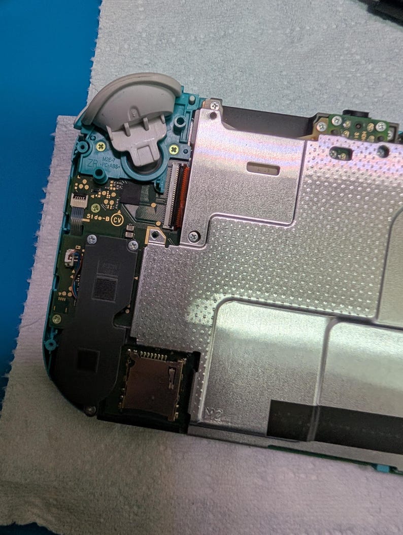 K&ouml;nnte beinhalten: Ein ge&ouml;ffnetes teal- und grau-farbenes Handheld-Spielger&auml;t, das interne Komponenten zeigt. Der obere linke Eckbereich des Ger&auml;ts zeigt einen abgenommenen grauen Joystick. Verschiedene Leiterplatten, Metallplatten und Anschl&uuml;sse sind sichtbar, was auf Reparatur oder Demontage hindeutet.