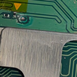 K&ouml;nnte beinhalten: Nahaufnahme einer Leiterplatte mit gr&uuml;nen und silbernen Komponenten. Die gr&uuml;ne Platine hat Kupferspuren und ein gelbes Dreieck. Ein silbernes Metallteil grenzt an die Platine.