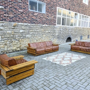 Könnte beinhalten: Eine Sitzgruppe im Freien mit einem braunen Ledersofa, einem passenden Loveseat und einem einzelnen Sessel. Die Möbel sind mit einem Holzrahmen konstruiert. Zwischen Sofa und Loveseat liegt ein gemusterter Teppich. Die Kulisse ist im Freien, vor einer Backstein- und Steinmauer.
