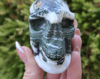 May include: A carved stone skull featuring a polished surface with distinct horizontal bands of deep forest green and opaque white mineral patterns. The skull has hollow eye sockets, a defined nasal cavity, and visible teeth detailing.