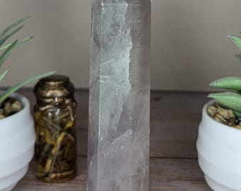 May include: A clear, hexagonal quartz crystal tower with a pointed top. The stone features internal fractures and cloudy inclusions throughout its structure, displaying a semi-transparent appearance with a smooth, polished surface finish.