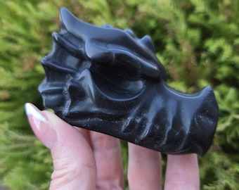 May include: A smooth, matte black stone carving shaped like a dragon head. The sculpture features detailed ridges along the snout, a prominent brow ridge, and a curved horn structure, showcasing a solid, dark, and uniform color throughout the piece.