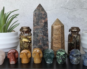 May include: A collection of eight small carved stone skulls in various colors including pink, red, yellow, brown, green, and blue, arranged in front of two tall stone obelisks and two glass jars filled with dried herbs and dark liquid.