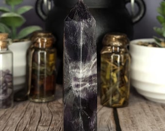 May include: A tall, hexagonal crystal point featuring deep purple and translucent white banded patterns. The polished stone has a pointed top and a flat base, displaying natural mineral inclusions and variations in color throughout its vertical structure.