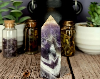 May include: A polished amethyst crystal tower featuring distinct chevron patterns of deep purple, white, and translucent quartz. The hexagonal point displays a natural color gradient from a light golden tip to dark purple and white banded layers.
