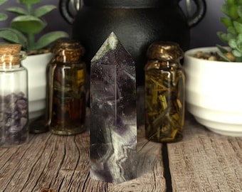 May include: A polished, hexagonal fluorite crystal tower featuring deep purple and white banded patterns. The stone stands upright, displaying a pointed termination and a smooth, translucent surface with natural color variations throughout the mineral.