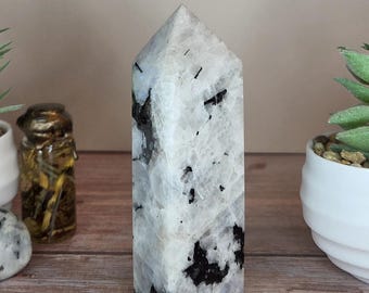 May include: A polished, six-sided crystal tower featuring a white and translucent base with prominent, irregular black mineral inclusions throughout the stone. The point is faceted into a pyramid shape, showcasing a smooth, reflective surface texture.