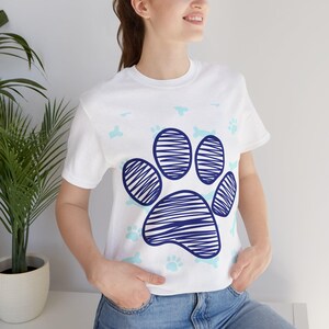 Pode incluir: Camiseta branca com um grande desenho de pegada azul. A pegada é feita de linhas azuis. Pequenas pegadas e ossos azuis estão espalhados pela camisa. A camisa é feita de um material macio.