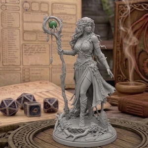 Könnte beinhalten: Graue Fantasy-Figur einer Frau, die einen Stab mit einer grünen Kugel hält. Die Figur steht auf einem runden Sockel, mit Würfeln und einem Spielbrett im Hintergrund. Ein geschnitztes Buch mit einem Drachen ist ebenfalls zu sehen.